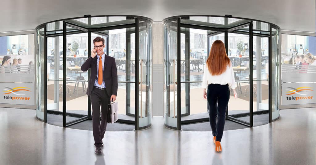 A businessman and a businesswoman use the revolving door of a modern office building. The man is wearing a suit and an orange tie, talking on his phone and holding a briefcase. The woman is wearing a white blouse and dark trousers, and is leaving the building. In the background, there are offices with glass walls and the "telepower" logo can be seen.