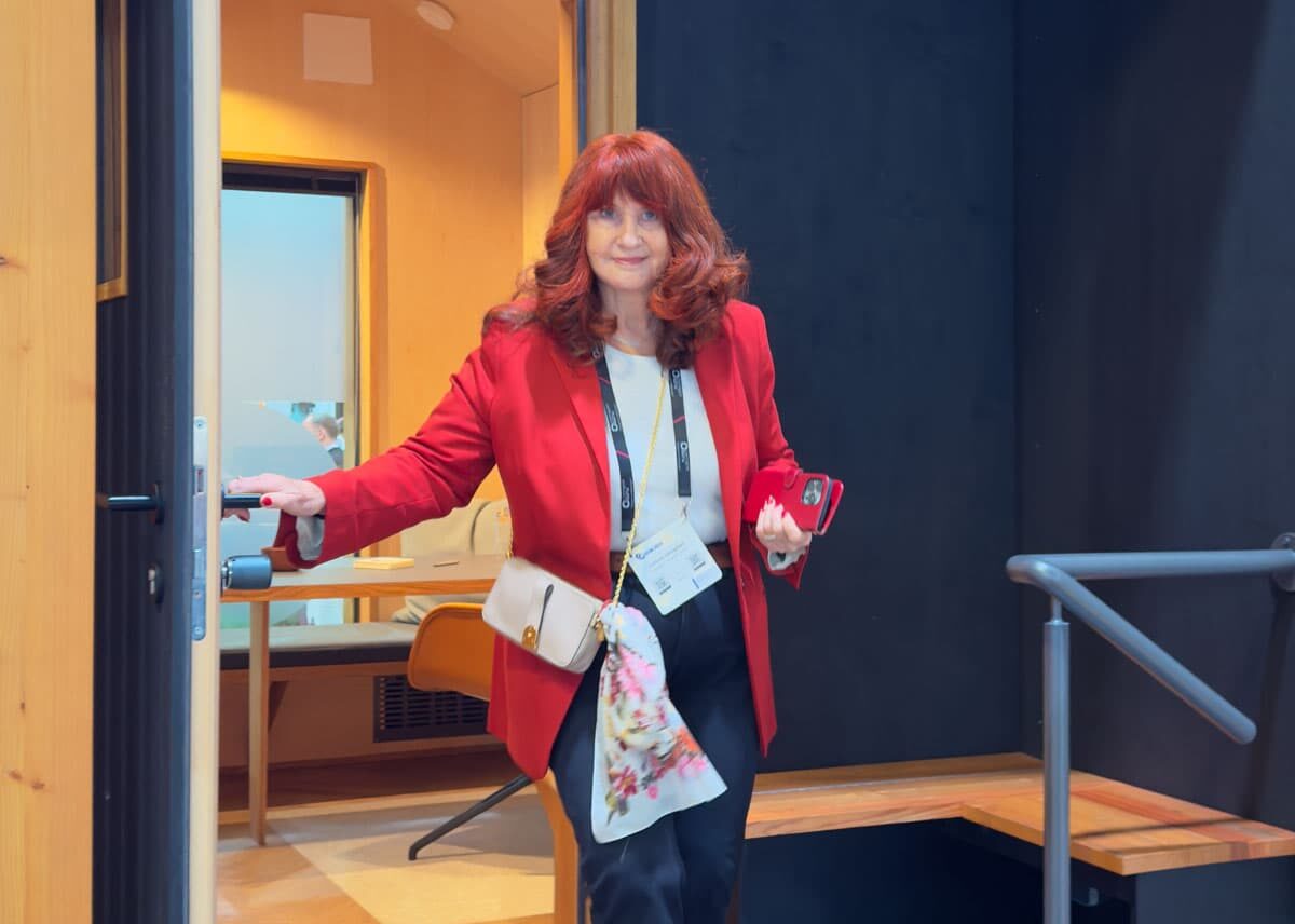Frau mit langen roten Haaren und rotem Blazer, die eine Tür öffnet und in einen Raum tritt.